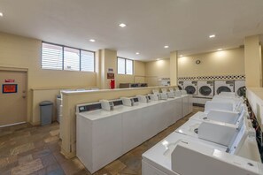 A laundry facility with multiple washing machines and dryers, a trash bin, and a sign indicating 'Workers Above'. - Royal Kuhio #1312 City Views (Honolulu)