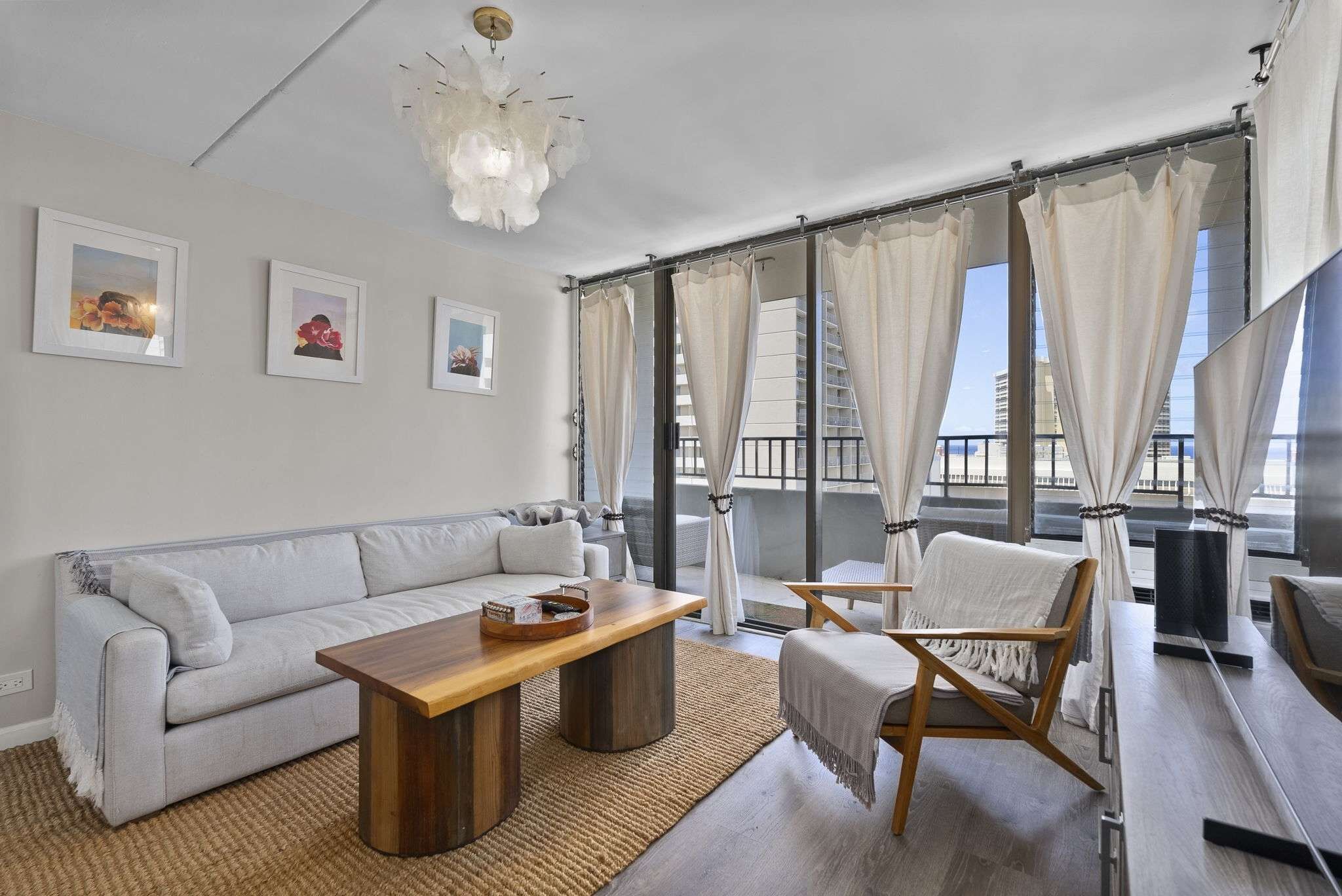 Ein modernes Wohnzimmer mit einer Couch, einem Holztisch und einem Sessel. Große Fenster mit Vorhängen, einer Deckenleuchte und gerahmten Bildern an der Wand.