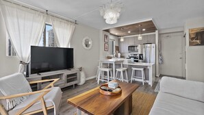A modern living room with a flat-screen TV, a wooden coffee table, and a sofa. There is a kitchen area with a refrigerator, microwave, and bar stools. - Royal Kuhio #1312 City Views (Honolulu)