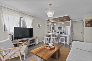 A modern living room with a flat-screen TV, a wooden coffee table, and a sofa. There is a kitchen area with a refrigerator, microwave, and bar stools. - Royal Kuhio #1312 City Views (Honolulu)