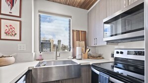 A modern kitchen with a stainless steel sink, wooden cabinets, a built-in oven, and a window with a city view. - Royal Kuhio #1312 City Views (Honolulu)