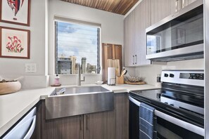 A modern kitchen with a stainless steel sink, wooden cabinets, a built-in oven, and a window with a city view. - Royal Kuhio #1312 City Views (Honolulu)