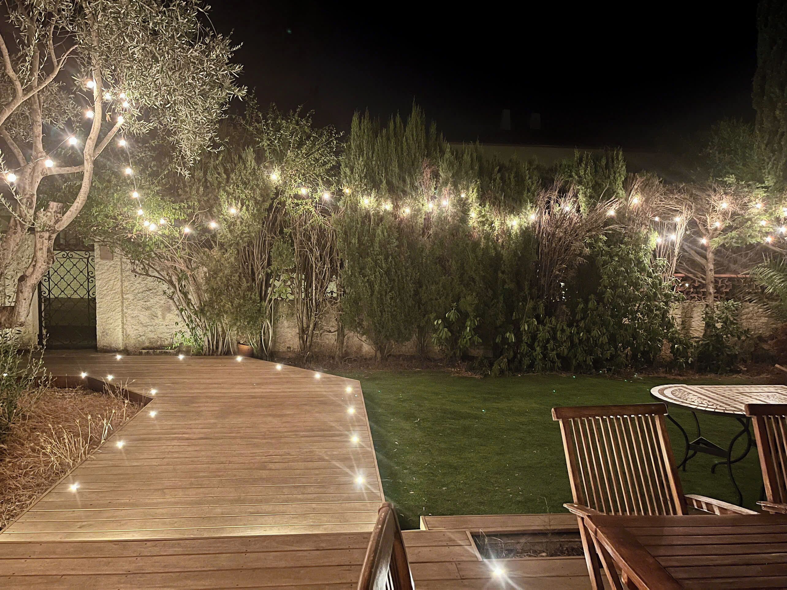 Een houten terras met lichtjes, een stenen muur en een gazon met een ronde tafel en stoelen.