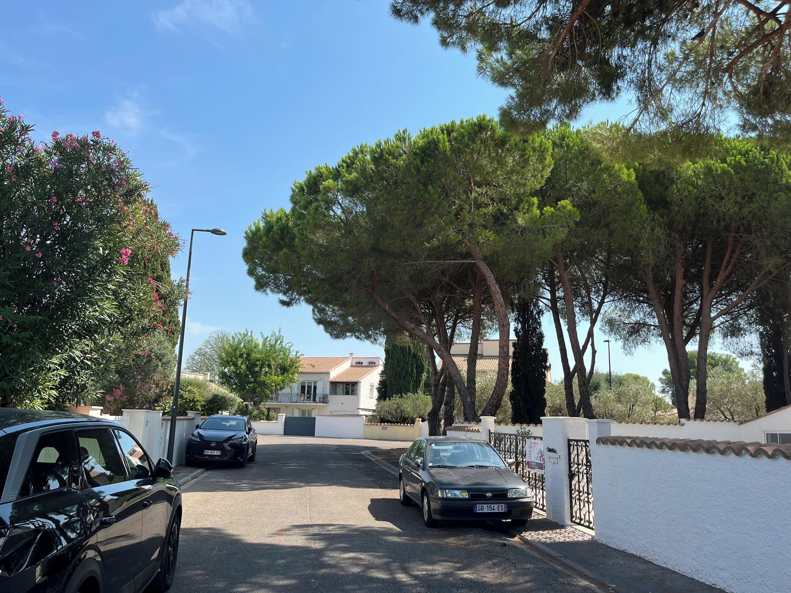 Una calle residencial con coches aparcados, árboles y un edificio blanco con tejado rojo.