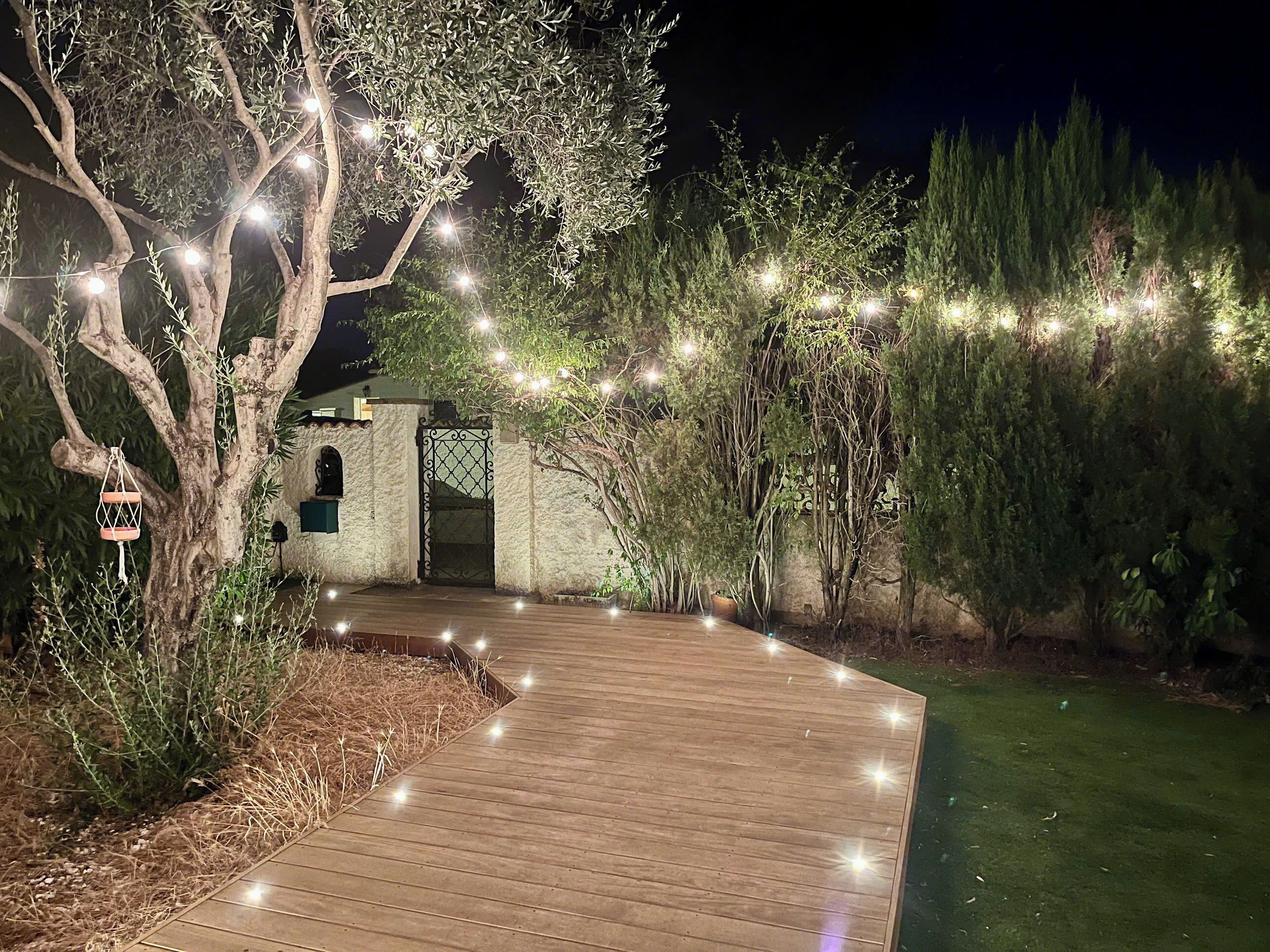 Een houten pad dat leidt naar een gebouw met een decoratieve poort, omgeven door groen en lichtjes.