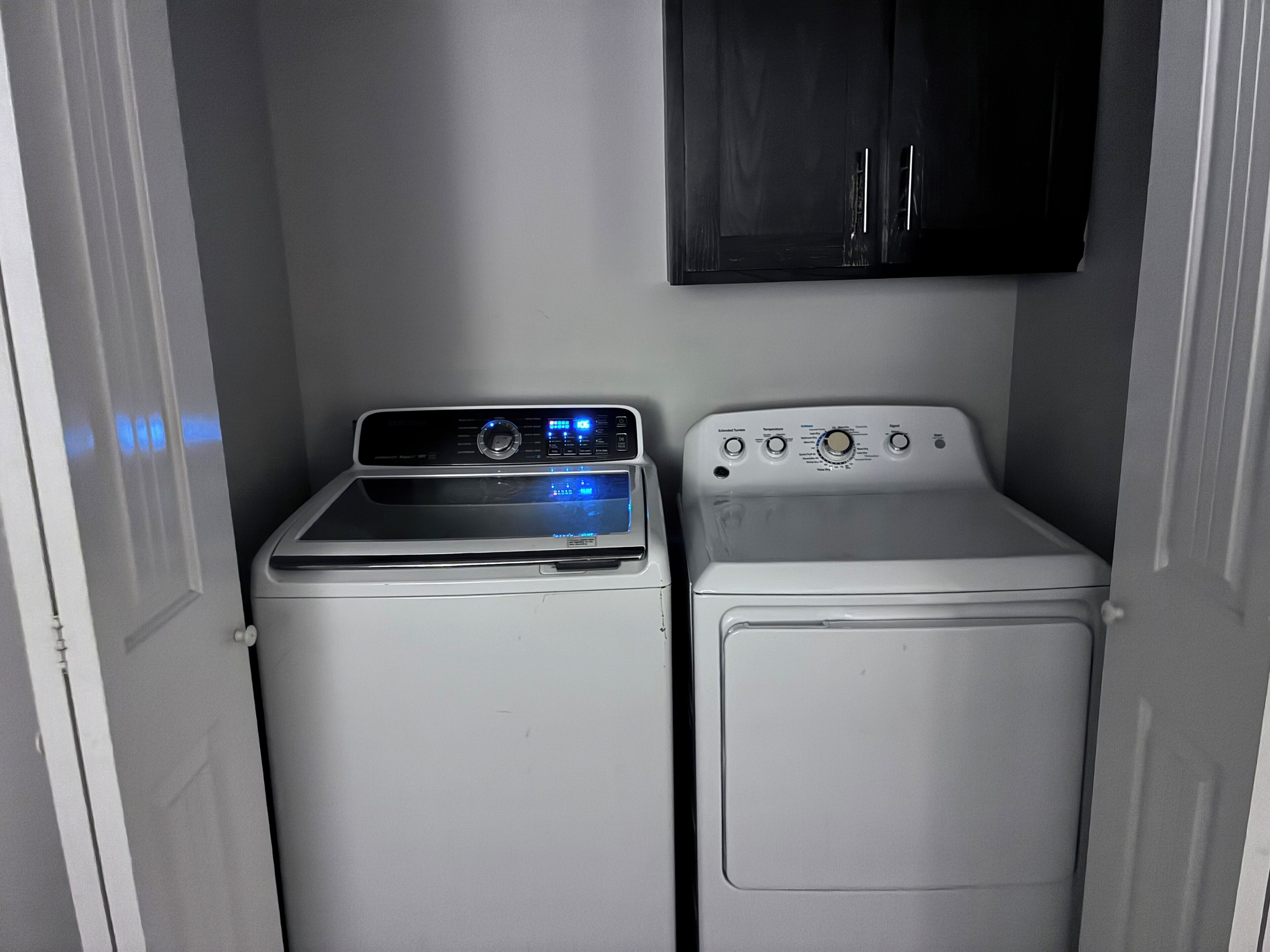 A compact laundry room with a front-loading washer and dryer, a dark cabinet above, and a door on the left.