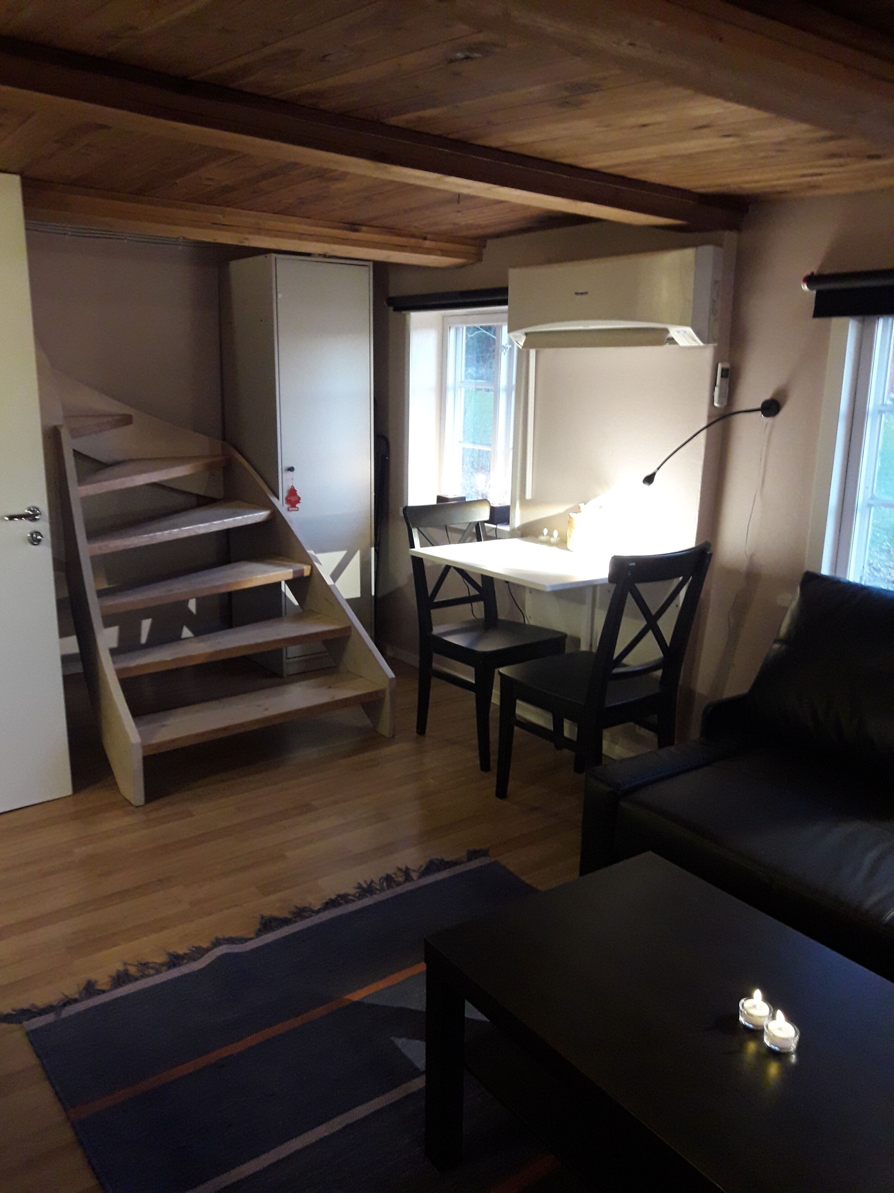 Ett rum med en trätrappa, ett skrivbord med en stol, en svart soffa och ett litet bord med ljus.