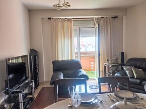 Ein Wohnzimmer mit einem schwarzen Ledersofa, einem Esstisch gedeckt mit Tellern und Besteck, einem Fernseher und einer Glastür, die auf eine Terrasse führt.