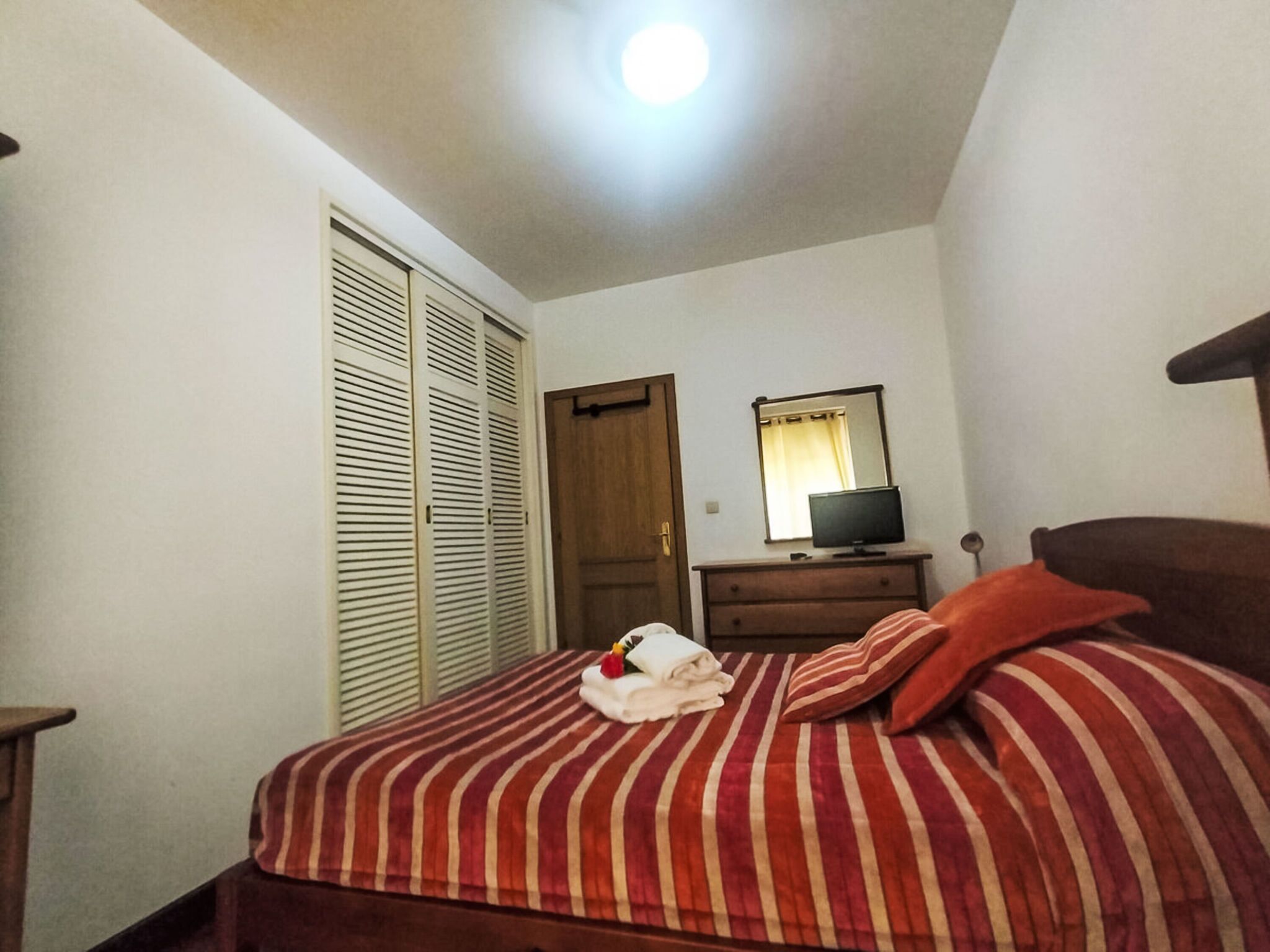 Une chambre à coucher avec un lit, un téléviseur posé sur une commode et une porte dotée d’un miroir.