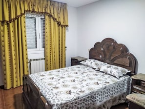 Quarto com cama, mesa de cabeceira, cabeceira de madeira e janela com cortinas.