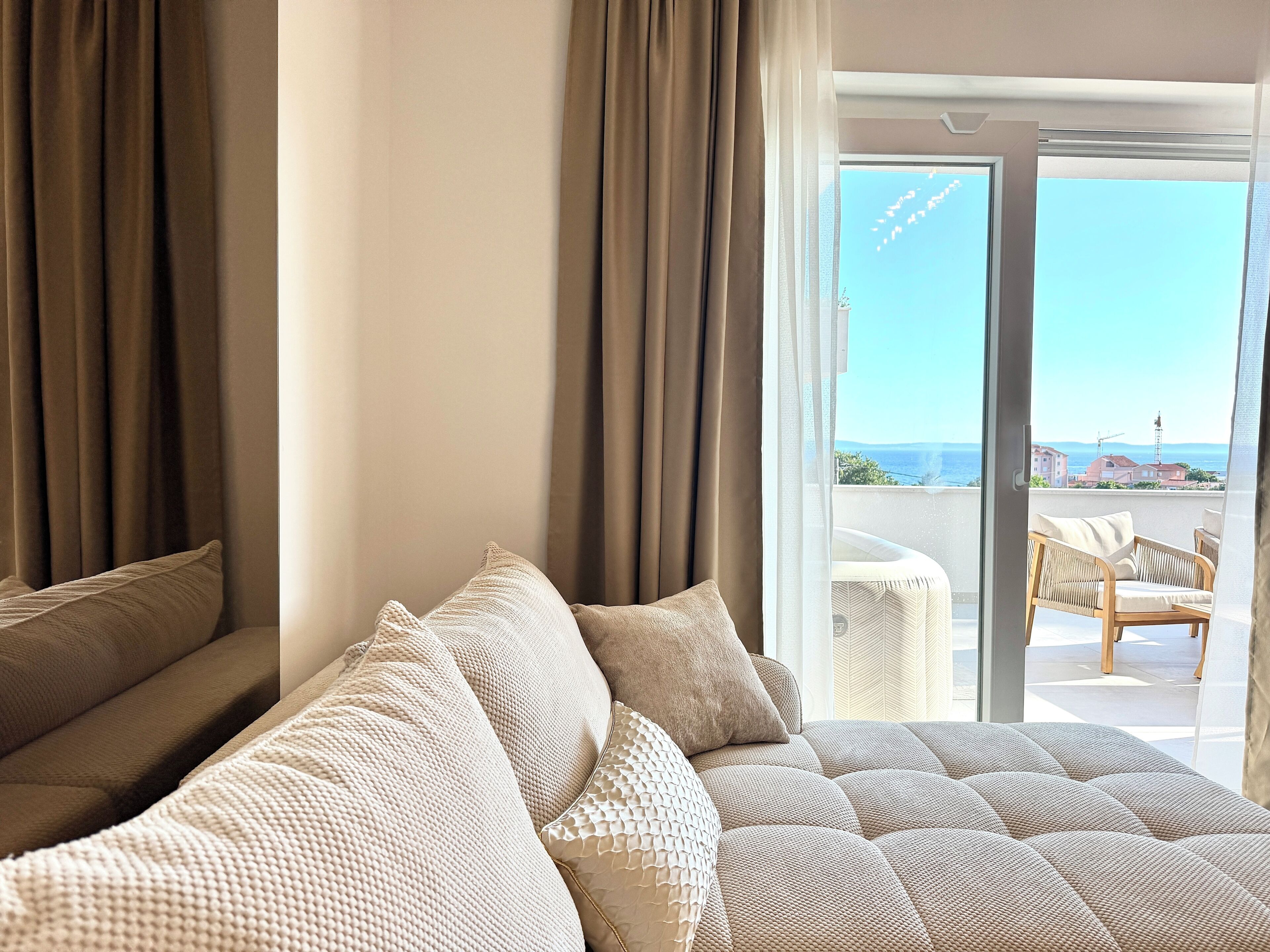 A hotel room with a large bed, multiple pillows, a view of the sea, and a balcony with outdoor furniture.