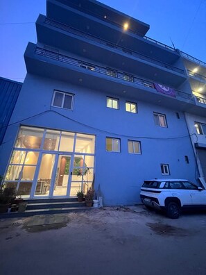 Un edificio de varios pisos con una entrada de vidrio amplia, un vehículo estacionado y un balcón visible.