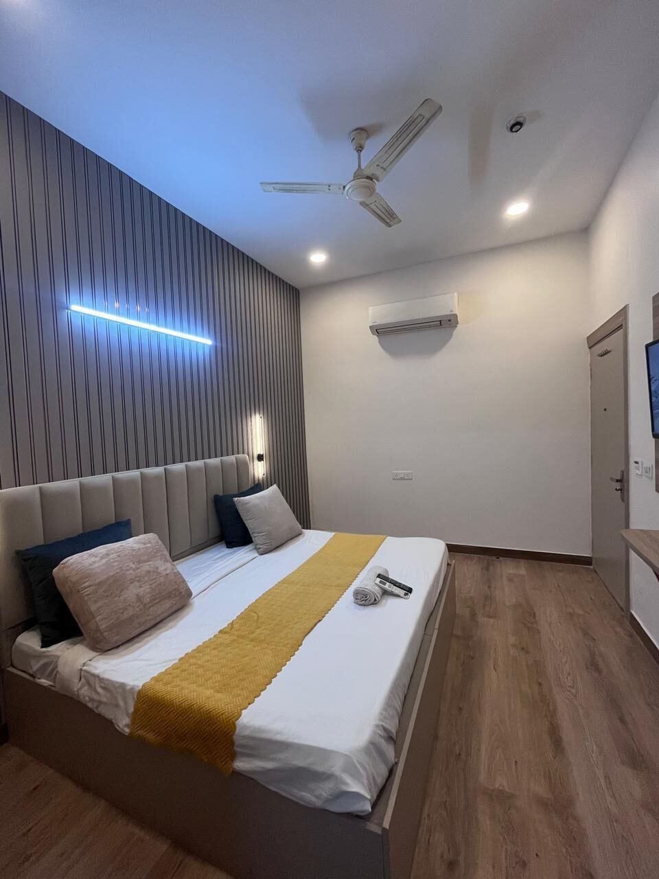 A modern bedroom with a bed, a ceiling fan, recessed lighting, an air conditioning unit, and a wooden floor.