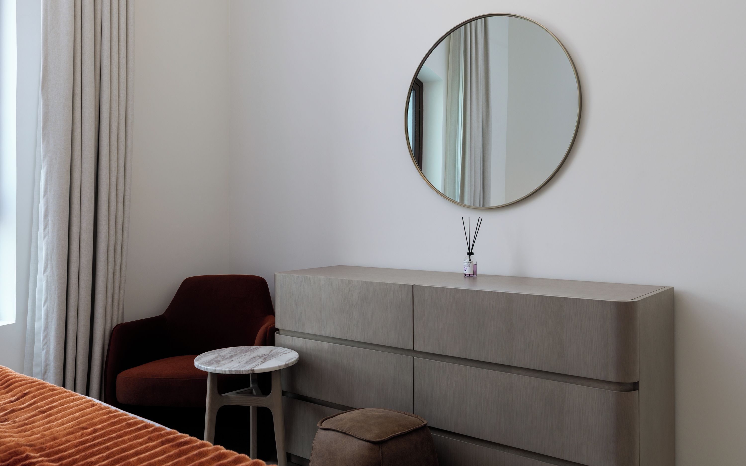 Ein Hotelzimmer mit einem Bett, einem roten Sessel, einem kleinen runden Tisch, einem grauen Schrank und einem runden Spiegel an der Wand.