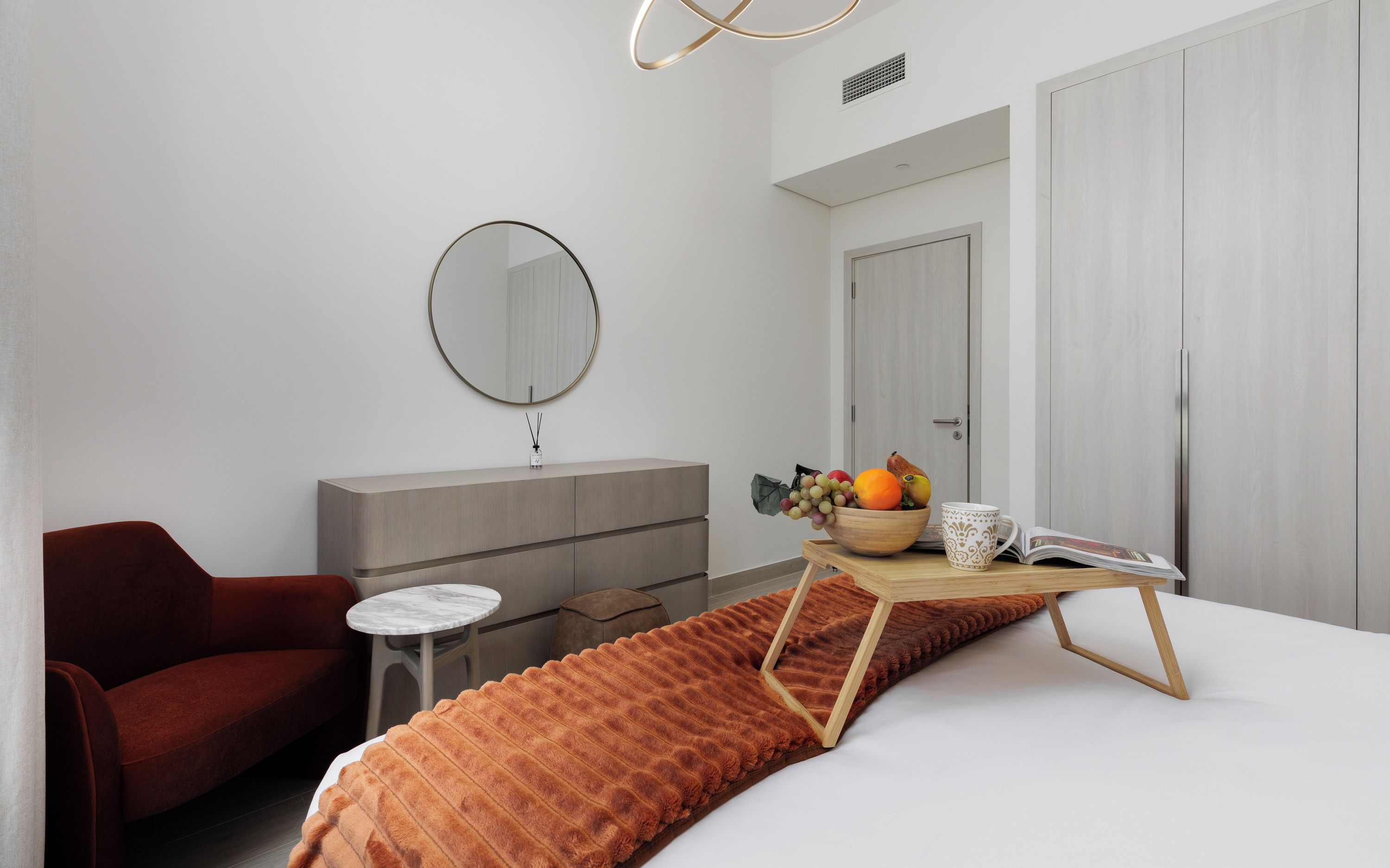 Ein modernes Schlafzimmer mit einem Bett, einem roten Sessel, einem kleinen Tisch mit Obstschale, einem Spiegel und einem Kleiderschrank.