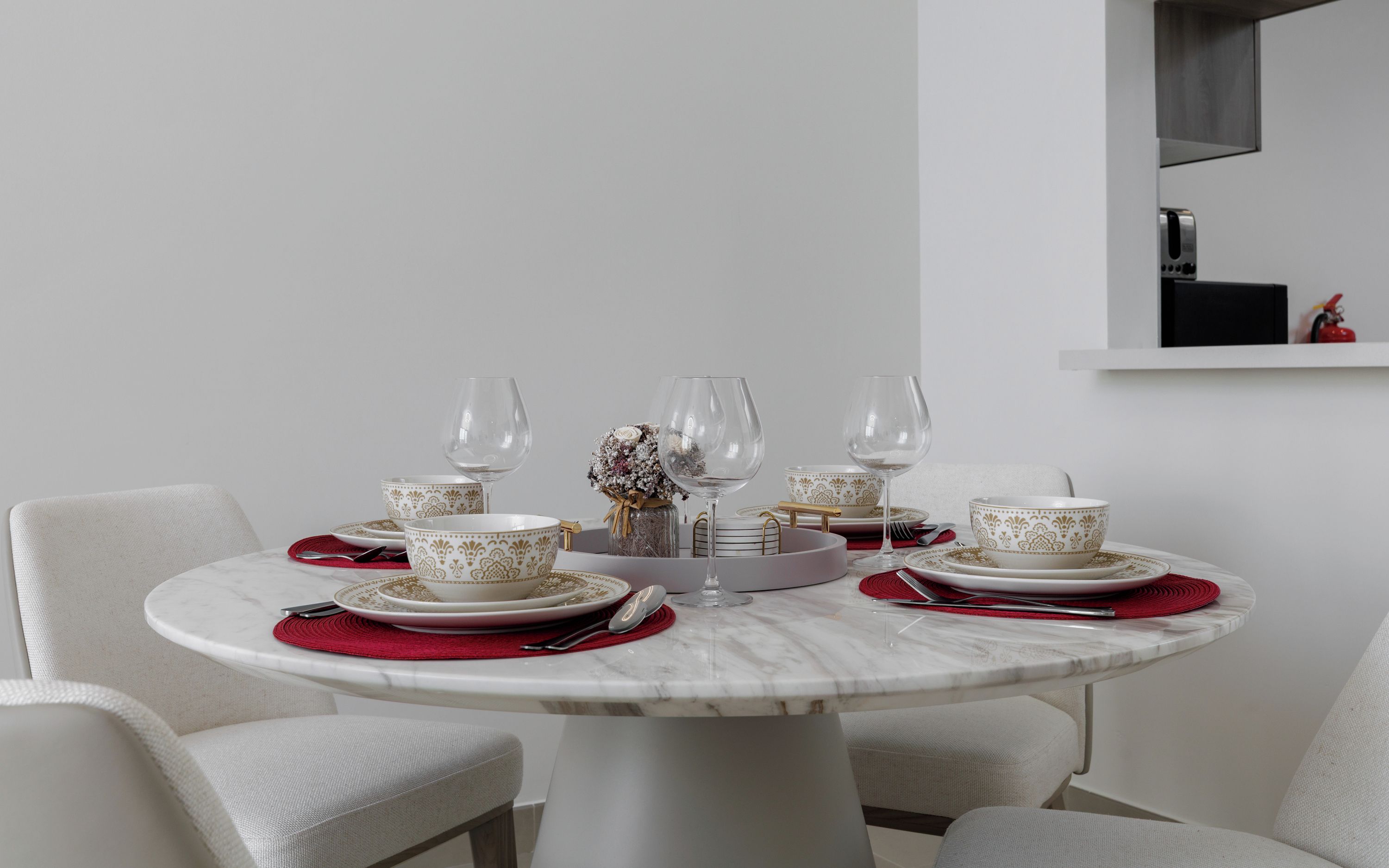 A round dining table with white chairs, a vase with dried flowers, and a small shelf with a toaster and a red object.