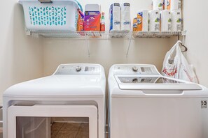 A laundry area with two front-loading washers and dryers, a shelf with laundry detergent, and a hanging bag. - Coastal Home on Hackler Golf Course Near CCU (Conway)