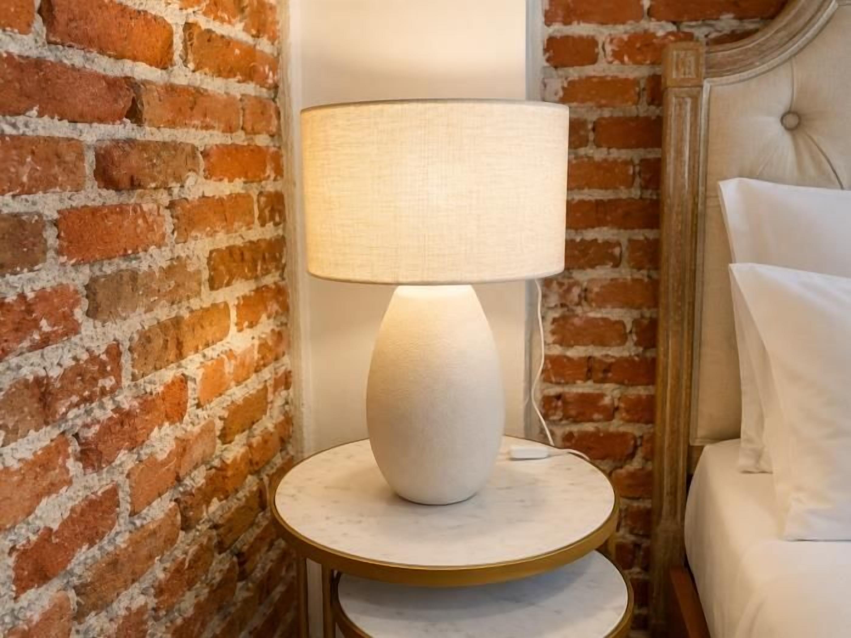 A lamp on a round table next to a brick wall and a bed with white bedding.