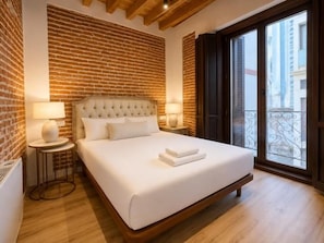 A hotel room with a brick wall, a large bed with white linens, two bedside lamps, and a view through a balcony door.