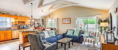 A spacious living area with a sofa, armchairs, and a coffee table. There is a kitchen area with wooden cabinets and a dining table.