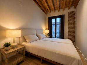 A double bed with a wooden headboard, bedside tables with lamps, a potted plant, and a view of a balcony with a brick wall.