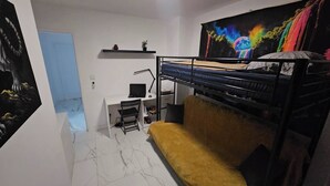Un dormitorio con literas, un escritorio con una computadora, una silla, un estante con objetos y un cuadro colorido en la pared.