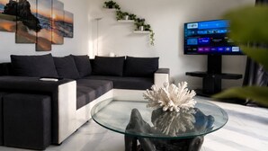 Una sala de estar moderna con un sofá seccional, una mesa de centro de vidrio con corales, un televisor de pantalla plana y un estante de pared con plantas.