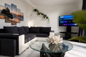 Ein modernes Wohnzimmer mit einem Ecksofa, einem Glastisch mit Korallen, einem Flachbildfernseher und einem an der Wand befestigten Regal mit Pflanzen.