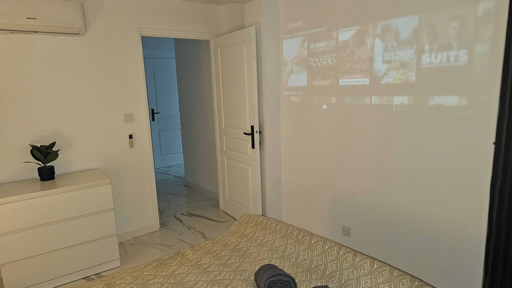 A bedroom with a bed, a dresser, a plant, a door, and a projection screen displaying images.