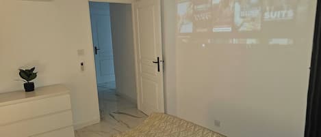 A bedroom with a bed, a dresser, a plant, a door, and a projection screen displaying images.