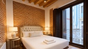 A hotel room with a brick wall, a large bed with white linens, two bedside lamps, and a view through a balcony door. - Juana I de Castilla - Apartamento Premium (Avila)