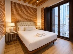 Een hotelkamer met een bakstenen muur, een groot bed met witte lakens, twee bedlampen en uitzicht door een balkondeur.