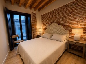 Ein Schlafzimmer mit einer Ziegelwand, einem Bett mit Fischgräten-Kopfteil, zwei Nachttischlampen, einem Stuhl und Blick durch eine Glastür.
