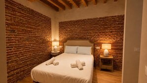 Ein Schlafzimmer mit Backsteinwand, Holzbalkendecke, einem Bett mit weißen Leinen, zwei Nachttischlampen und einem Nachttisch aus Holz.