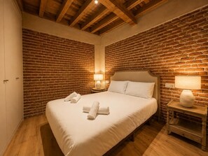 Habitación de hotel con pared de ladrillo, techo de madera, cama con ropa blanca y dos lámparas de noche.