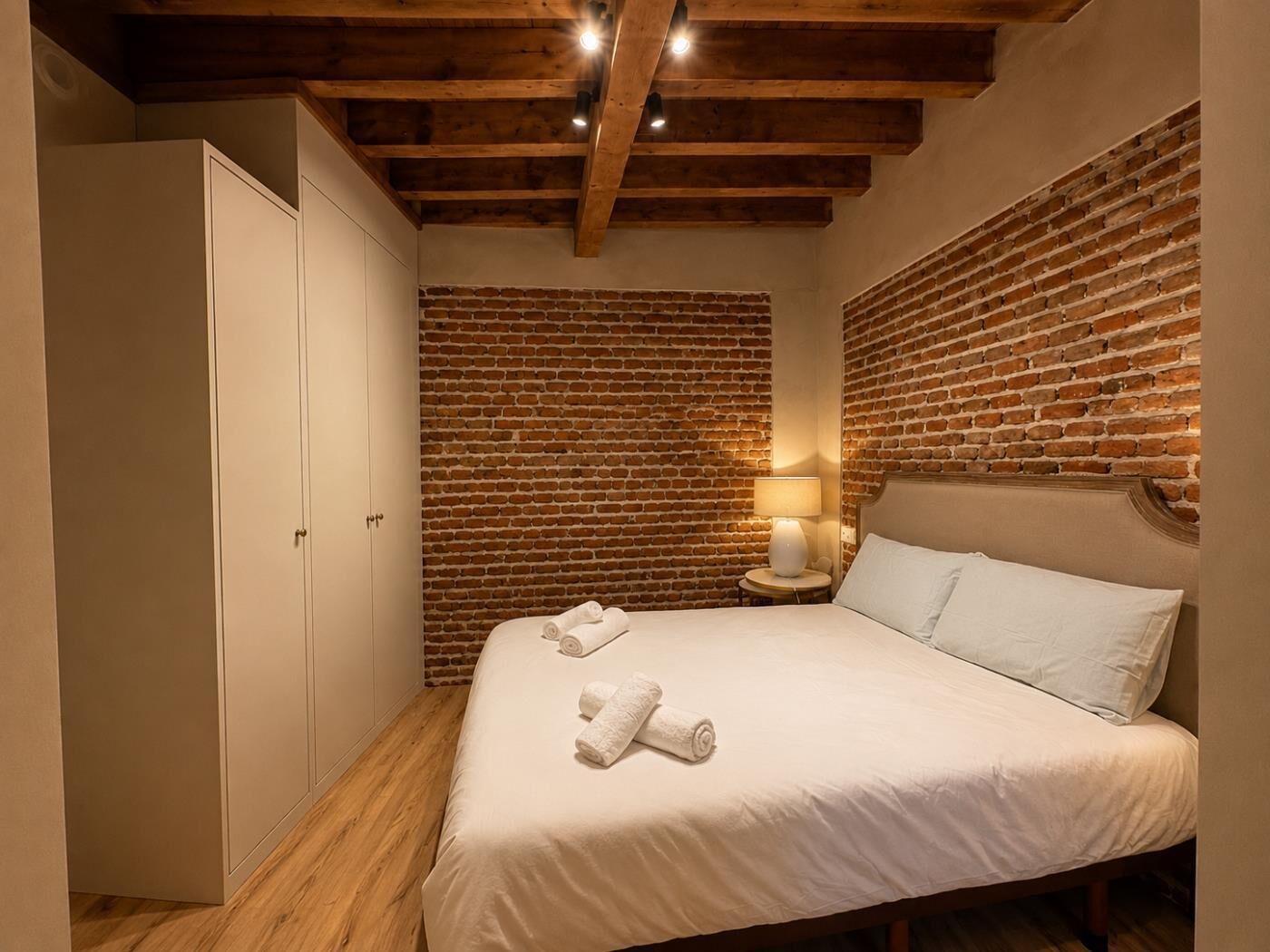 A bedroom with a brick wall, a bed with white linens, a nightstand with a lamp, and a wardrobe.