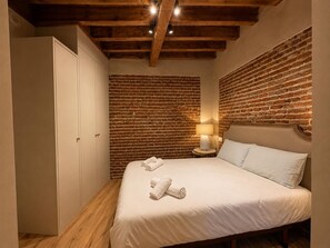 A bedroom with a brick wall, a bed with white linens, a nightstand with a lamp, and a wardrobe. - Berenguela de Castilla - Apartamento Deluxe (Avila)