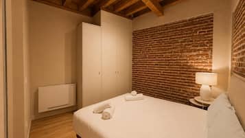 A bedroom with a brick wall, wooden ceiling beams, a bed with white linens, a bedside table with a lamp, and a wardrobe.