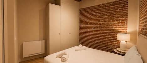 Un dormitorio con pared de ladrillo, vigas de madera en el techo, una cama con ropa de cama blanca, una mesita de noche con lámpara y un armario.