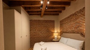A bedroom with a brick wall, a bed with white linens, a nightstand with a lamp, and a wardrobe. - Leonor de Aquitania - Apartamento Deluxe (Avila)