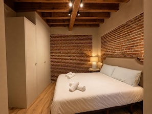A bedroom with a brick wall, a bed with white linens, a nightstand with a lamp, and a wardrobe. - Leonor de Aquitania - Apartamento Deluxe (Avila)
