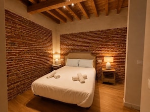 A bedroom with a brick wall, wooden ceiling beams, a bed with white linens, two bedside lamps, and a wooden nightstand. - Leonor de Aquitania - Apartamento Deluxe (Avila)