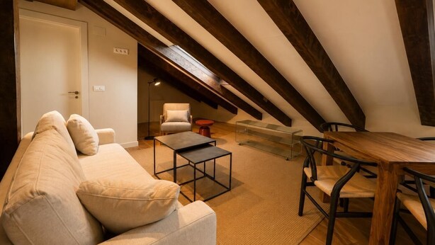 An attic room with a high ceiling, exposed wooden beams, a sofa, a coffee table, a chair, and a dining table. - Santa Teresa de Jesús - Apartamento Deluxe (Avila)