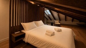 A bedroom with a sloped ceiling, a bed with white linens, a chair, and a small table. - Santa Teresa de Jesús - Apartamento Deluxe (Avila)