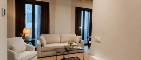 A living room with a sofa, armchair, and coffee table. There are windows with curtains, a lamp, and a plant.