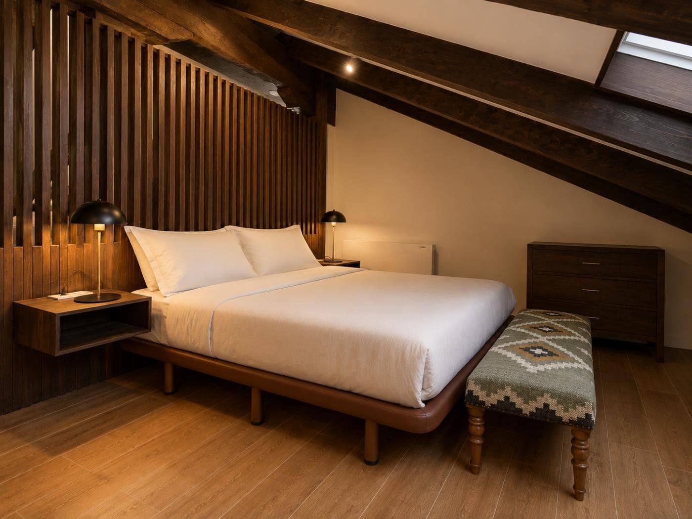 A bedroom with a sloped ceiling, a bed with white bedding, two bedside lamps, a wooden nightstand, a bench with a patterned cushion, and a wooden dresser.