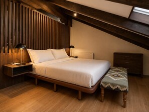 A bedroom with a sloped ceiling, a bed with white bedding, two bedside lamps, a wooden nightstand, a bench with a patterned cushion, and a wooden dresser.