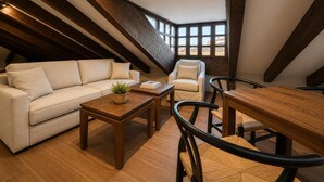 A loft-style room with a sloped ceiling, wooden beams, a sofa, a chair, a table, and a view through large windows. - María de Molina - Apartamento Deluxe (Avila)