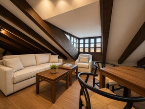 Een loft-achtige kamer met een schuine kap, houten balken, een bank, een stoel, een tafel en uitzicht door grote ramen.