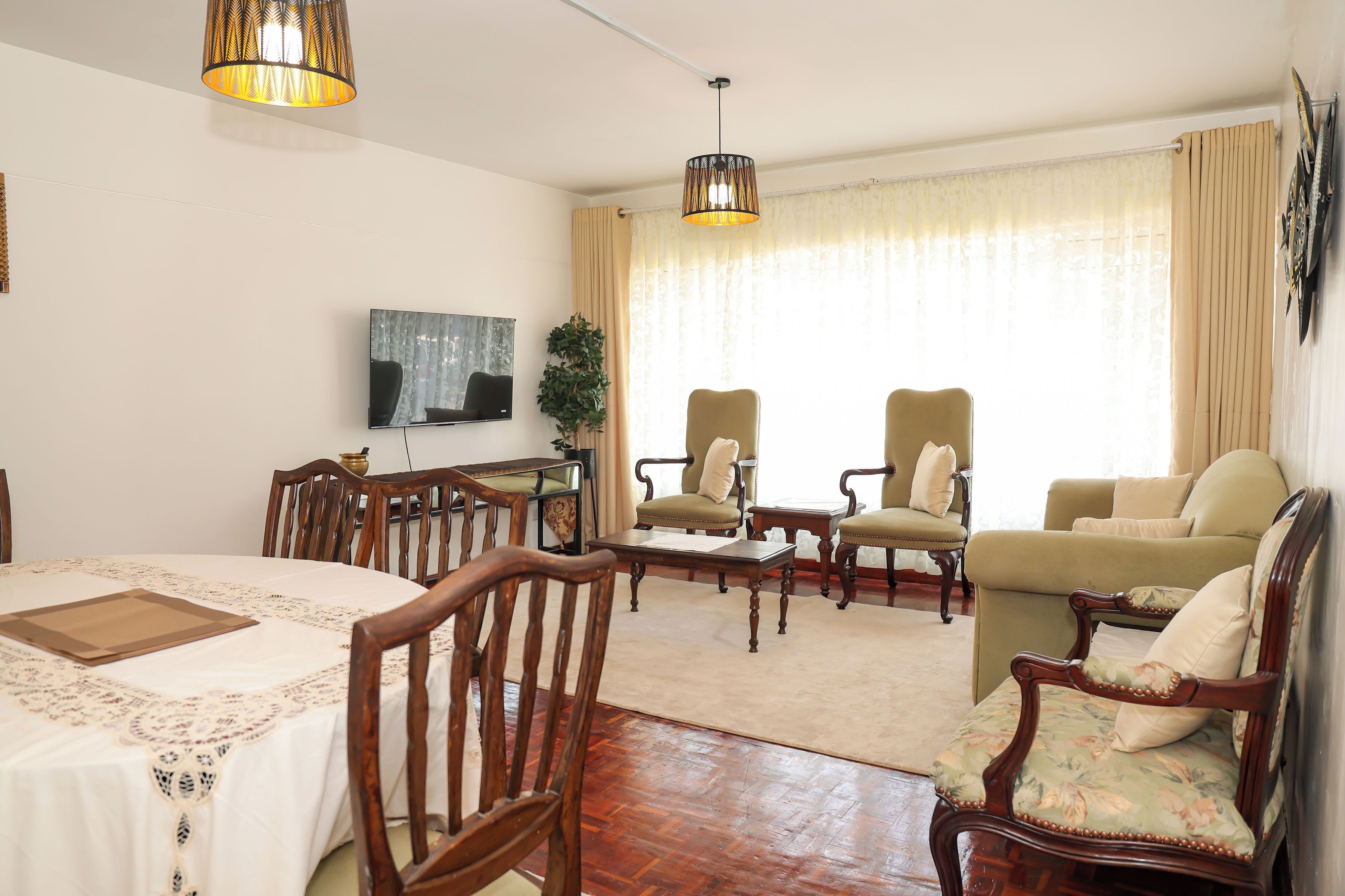 A living room with a dining area, wooden furniture, a flat-screen TV, and a large window with curtains.